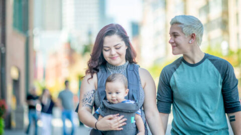 A queer couple is walking in an urban area along with their baby in a carrier.