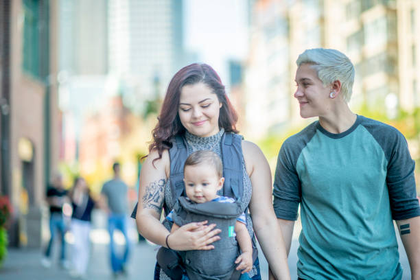 A queer couple is walking in an urban area along with their baby in a carrier.